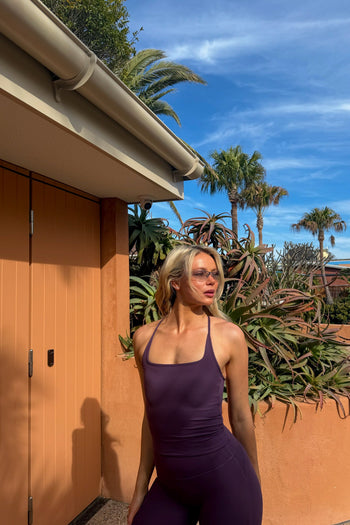 A woman wearing a dark purple activewear set, tank and flares, stands outdoors next to a terracotta-colored wall and door, with palm trees and a blue sky in the background.