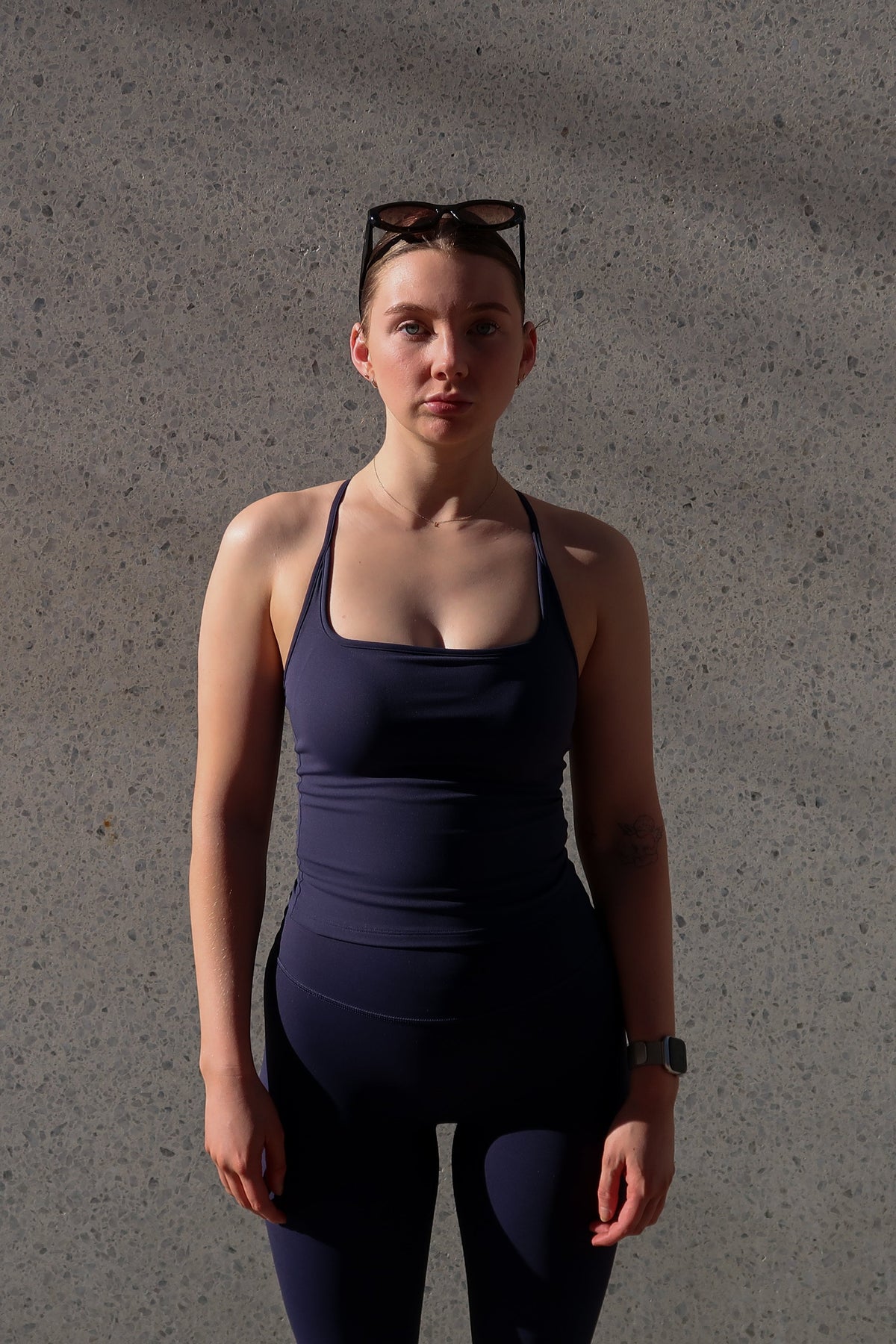 A woman stands against a textured concrete wall wearing a dark navy blue athletic tank top and matching leggings, with sunglasses resting on her head.