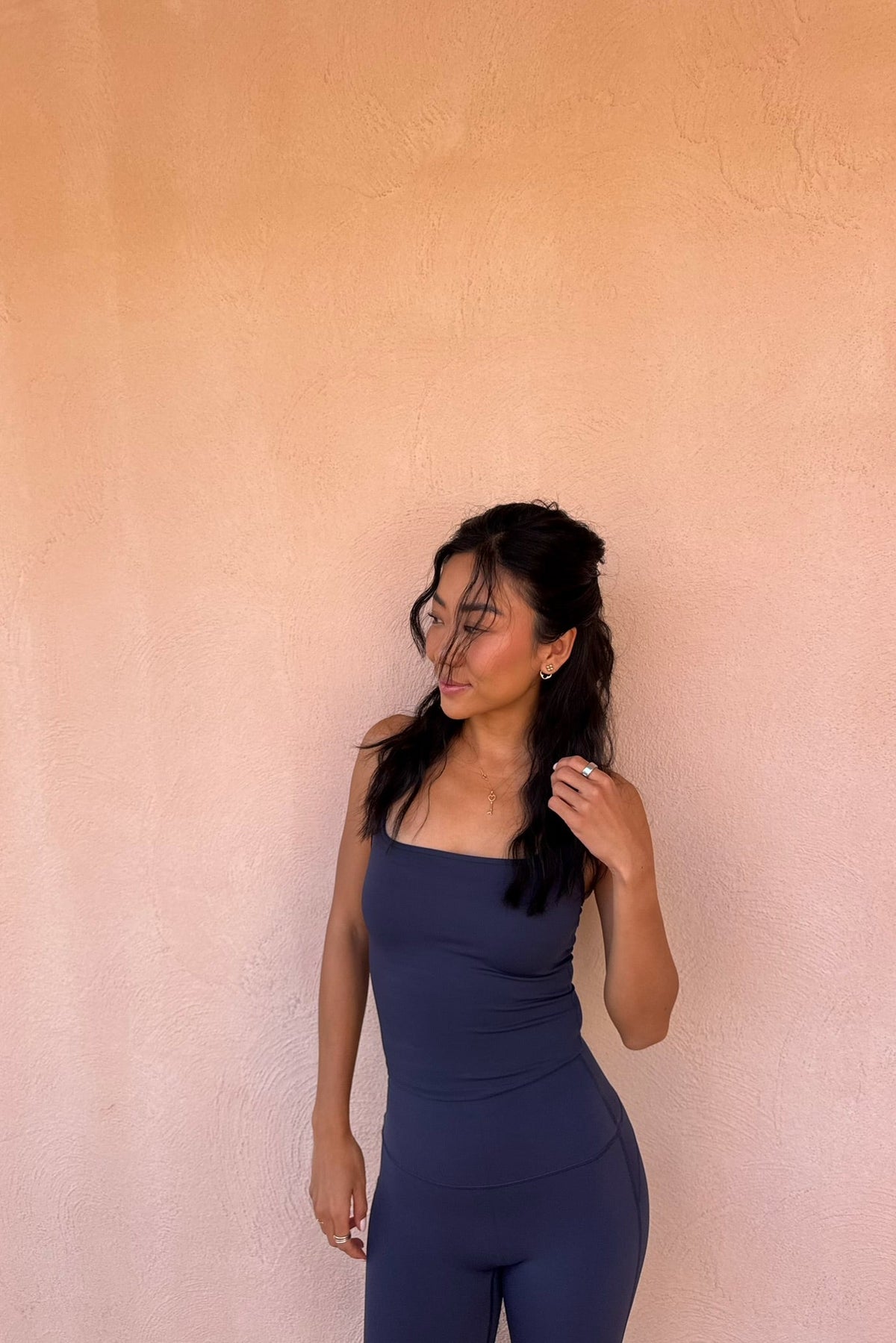 A woman wearing a dark slate blue ribbed tank top and matching leggings stands against a textured peach-colored stucco wall.