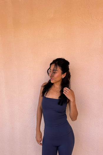 A woman wearing a dark slate blue ribbed tank top and matching leggings stands against a textured peach-colored stucco wall.